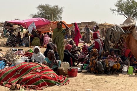 People surrounded by belongings sit on the ground, some under flimsy sunshades make of sheets and sticks, others in the sun.