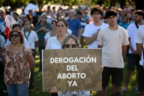 People standing in rows looking like they are singing an anthem while an older woman at the front holds a sign saying: 'Derogación del Aborto ya'